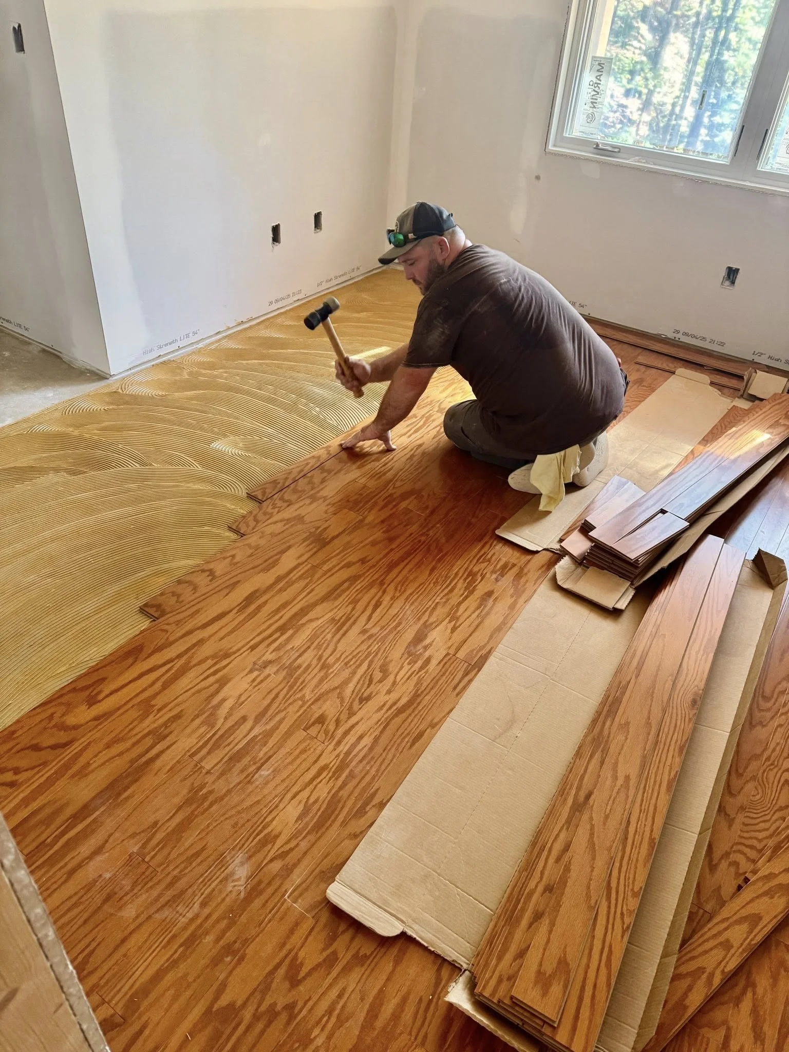 Matt Smith installing hardwood flooring in a room with a large window. He is kneeling and using a mallet to secure planks while arranging multiple wood pieces.