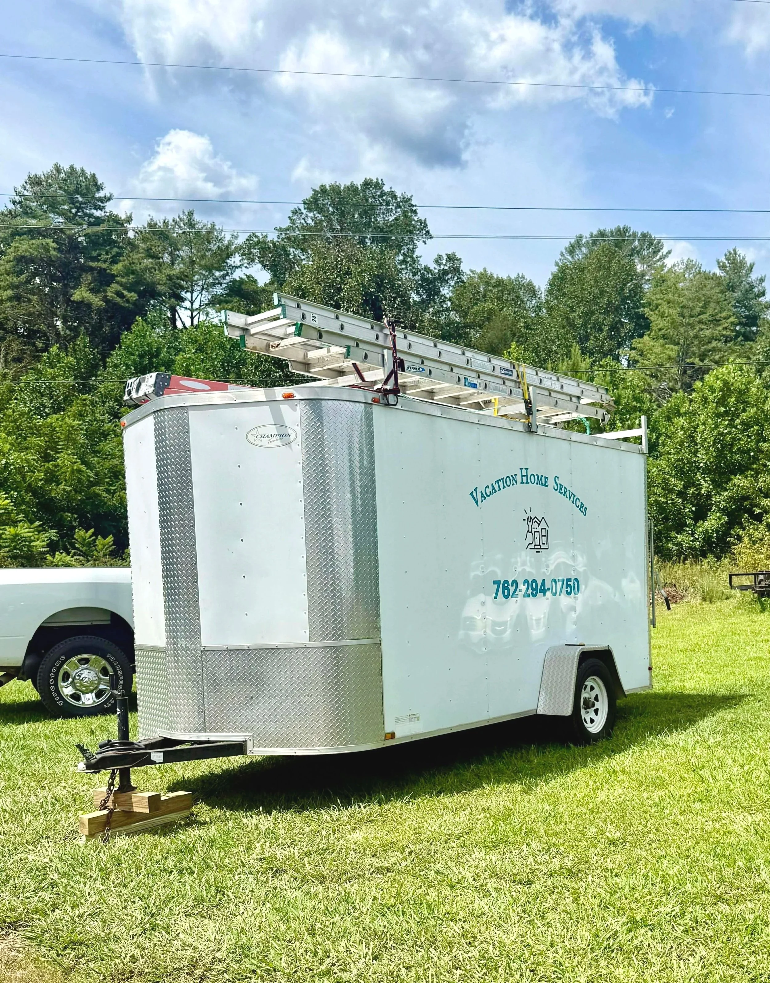 White mobile service trailer with a ladder on top, parked on grass in a green outdoor area with trees and partly cloudy sky in the background.