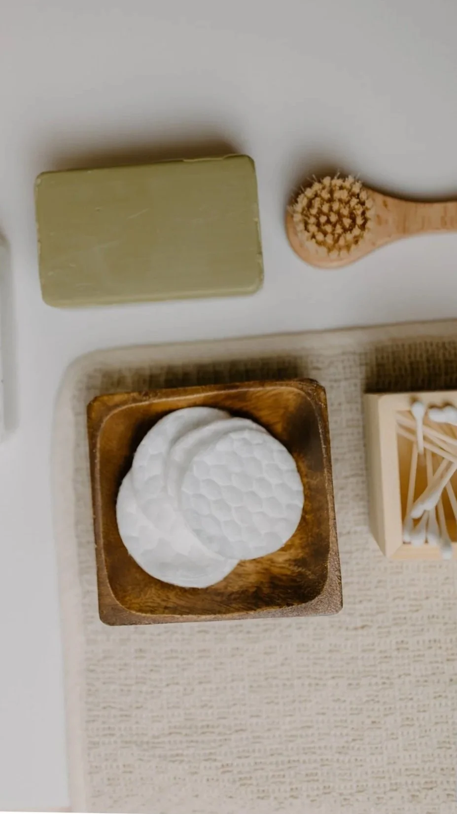 A wooden bowl with white honeycomb-patterned cotton pads, a small wooden box with cotton swabs, a green soap bar, and a wooden facial massager with rounded bristles on a white surface.