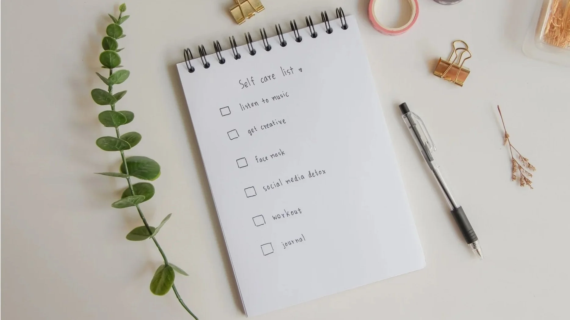A white notepad titled 'Self care list' with checked boxes for tasks such as listening to music, getting creative, face mask, social media detox, workout, and journal. The notepad is on a light-colored desk alongside a black pen, green plant branch, gold binder clip, pink tape, dried flower, and a container of toothpicks.