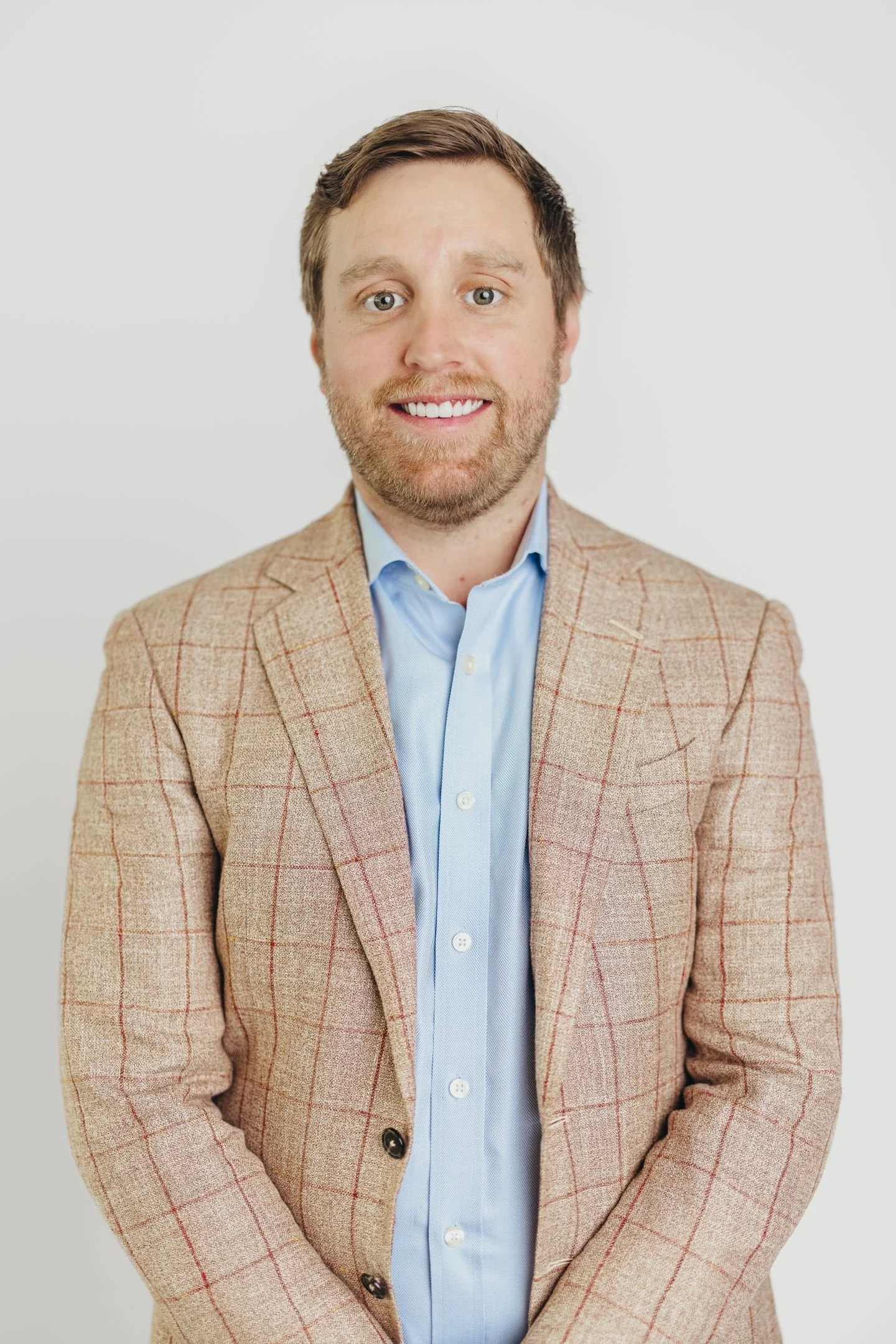 A man in a beige plaid blazer and light blue dress shirt, smiling against a plain white background.