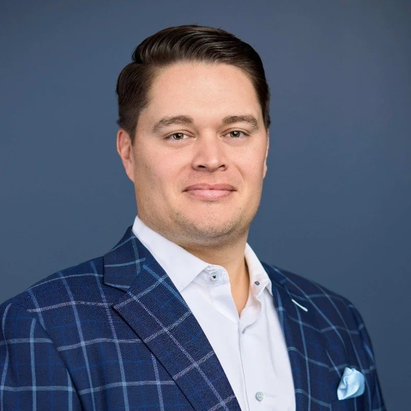A professional headshot of a man with fair skin, short dark hair, wearing a navy blue plaid blazer, white dress shirt, and a light blue pocket square, against a gray background.