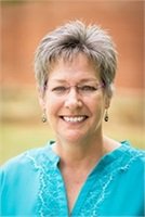 Portrait of a smiling woman with short gray hair wearing glasses and a turquoise blouse, against a yellow background.