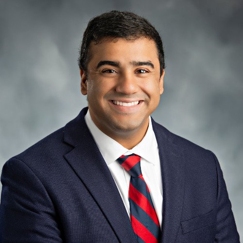 Professional headshot of a man with short dark hair, wearing a navy blue suit, white shirt, and striped red and blue tie, smiling against a gray background.