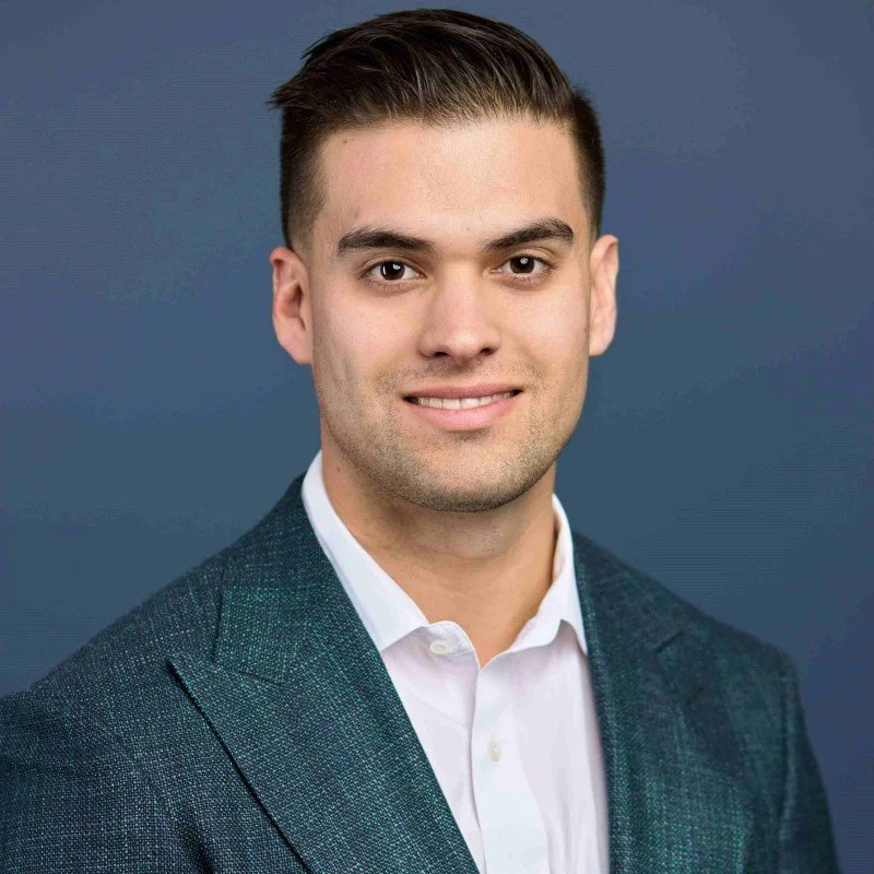 Professional headshot of a young man in a white shirt and dark blazer, smiling against a blue background.