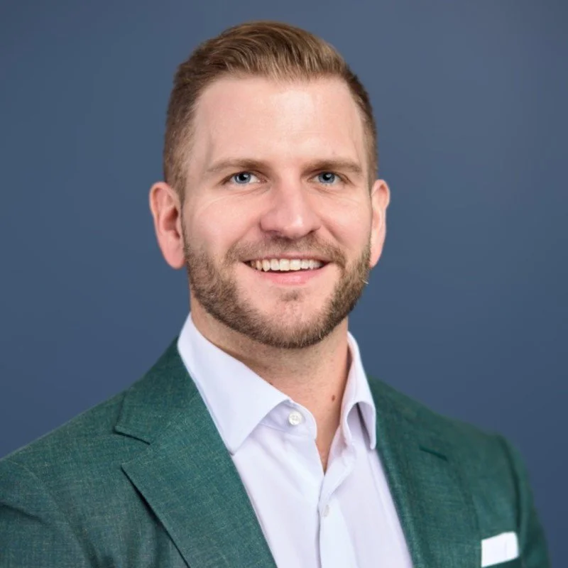 Headshot of a smiling man with short brown hair, blue eyes, and a beard, wearing a white shirt and green blazer against a blue background.