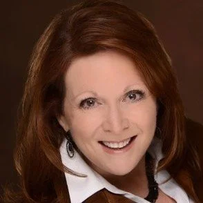 A smiling woman with shoulder-length red hair wearing a white collared shirt and dark earrings, against a brown background.