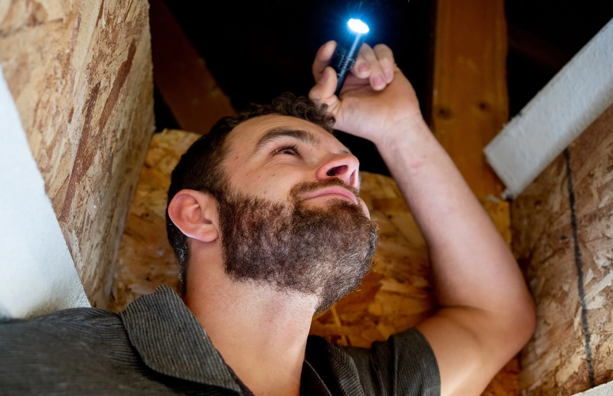 Man with a beard lying on his back inspecting a ceiling with a flashlight.