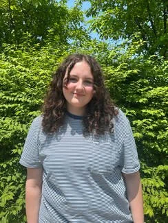 Young woman with curly dark hair wearing a gray and white striped t-shirt outdoors with green trees and blue sky in the background.