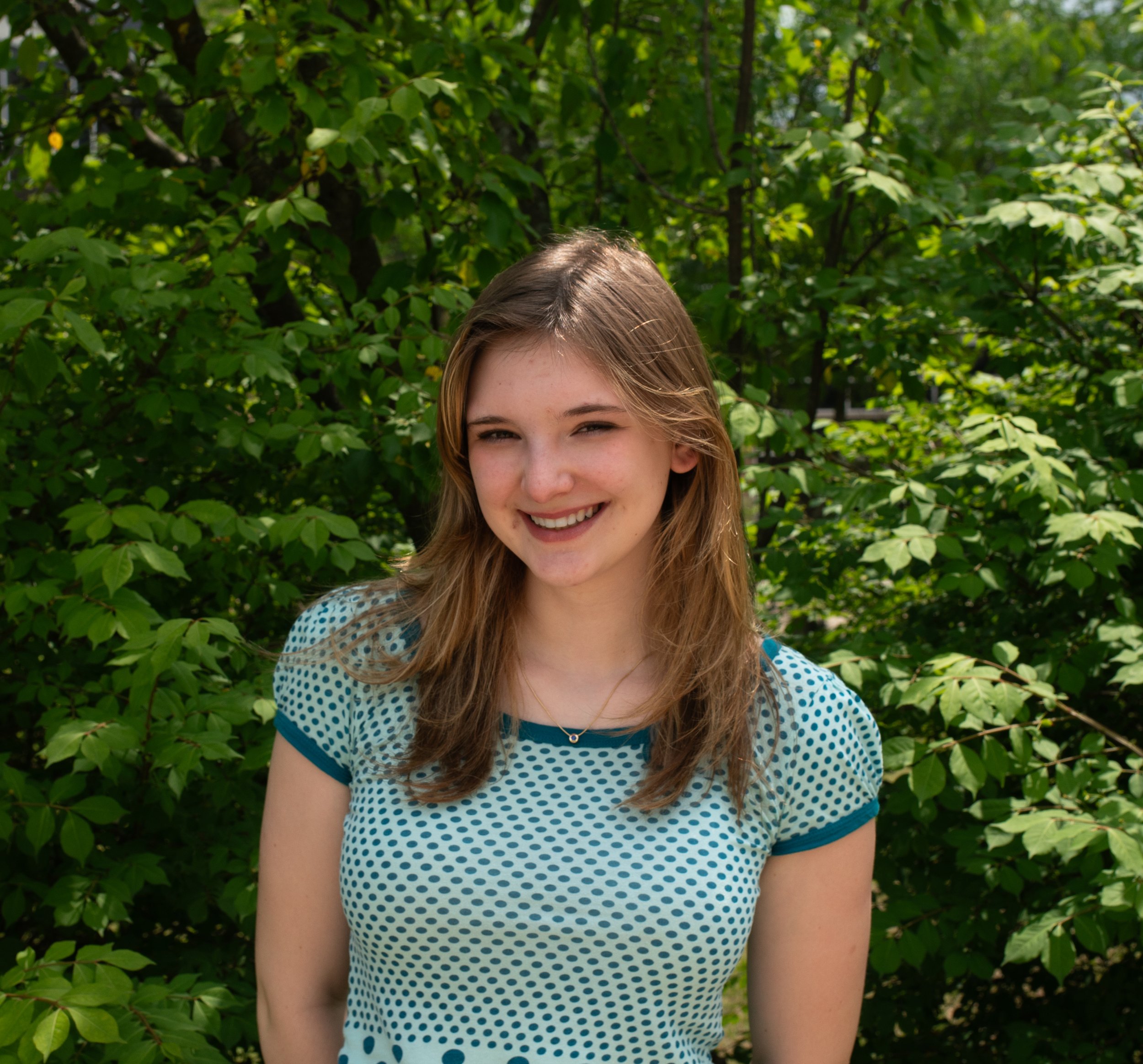 A young woman standing outdoors in front of lush green trees, smiling at the camera.