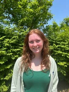 A young woman with red hair smiling outdoors, standing in front of green trees and a blue sky.