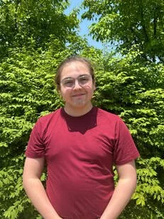 Person with glasses wearing a maroon t-shirt smiling outdoors with green trees and bushes in the background.