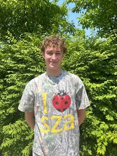 Young man standing outdoors in front of green trees, wearing a gray tie-dye T-shirt with a ladybug graphic and yellow text.