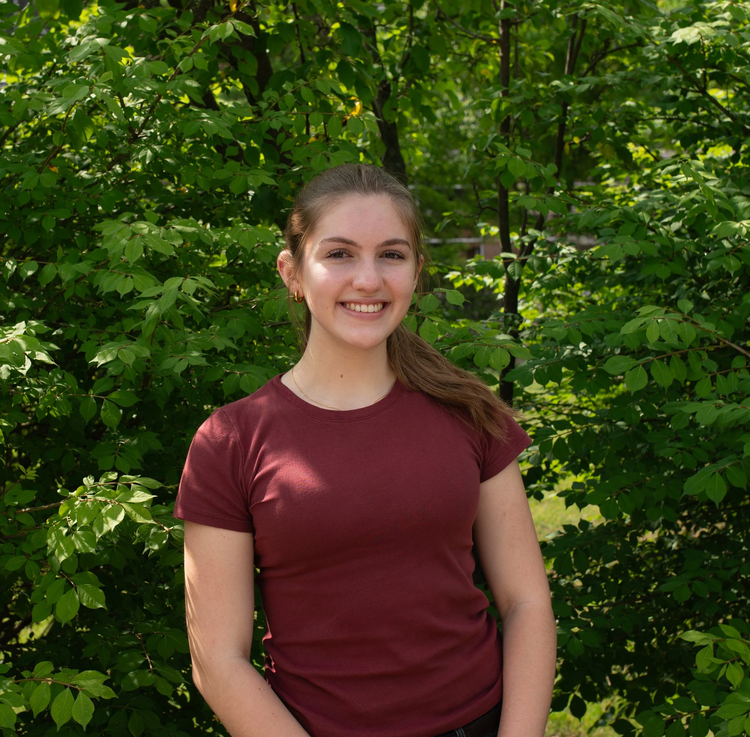 Young woman smiling outdoors with green trees and blue sky in the background.