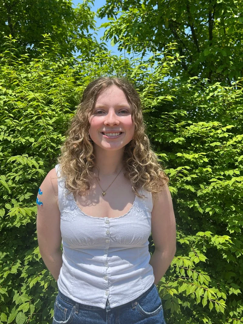 A young woman with curly blonde hair smiling outdoors in front of green foliage on a sunny day.