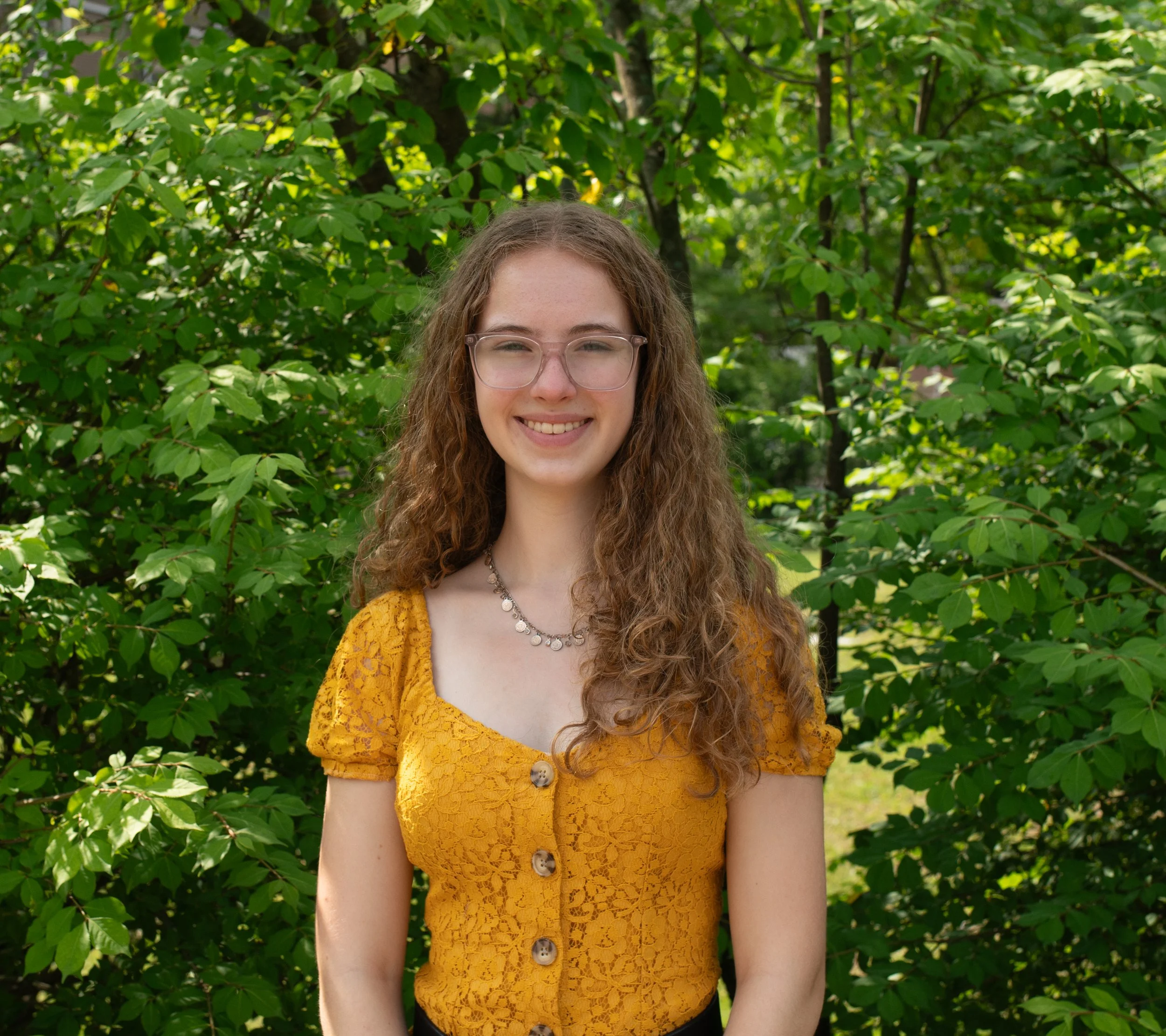 A young woman with curly blonde hair smiling outdoors in front of green foliage on a sunny day.