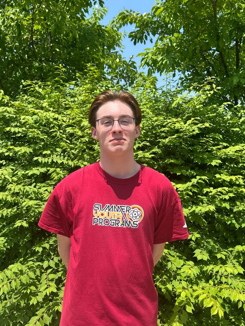 A young man with brown hair and glasses standing outdoors in front of green leafy bushes and trees on a sunny day.