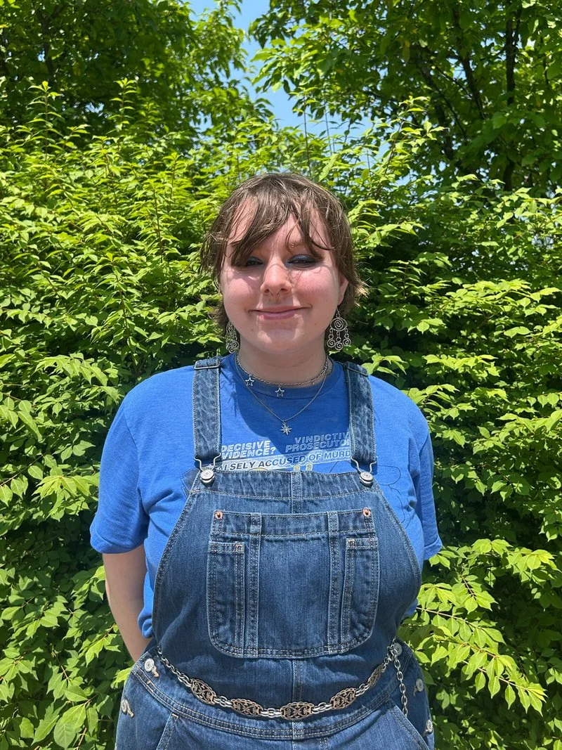 A young woman with short brown hair and blue eyes stands outdoors in front of lush green foliage, smiling with closed eyes. She is wearing a blue t-shirt and denim overalls with a chain belt, along with earrings and layered necklaces.