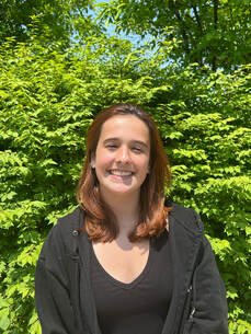 Young woman smiling outdoors with green trees and blue sky in the background.
