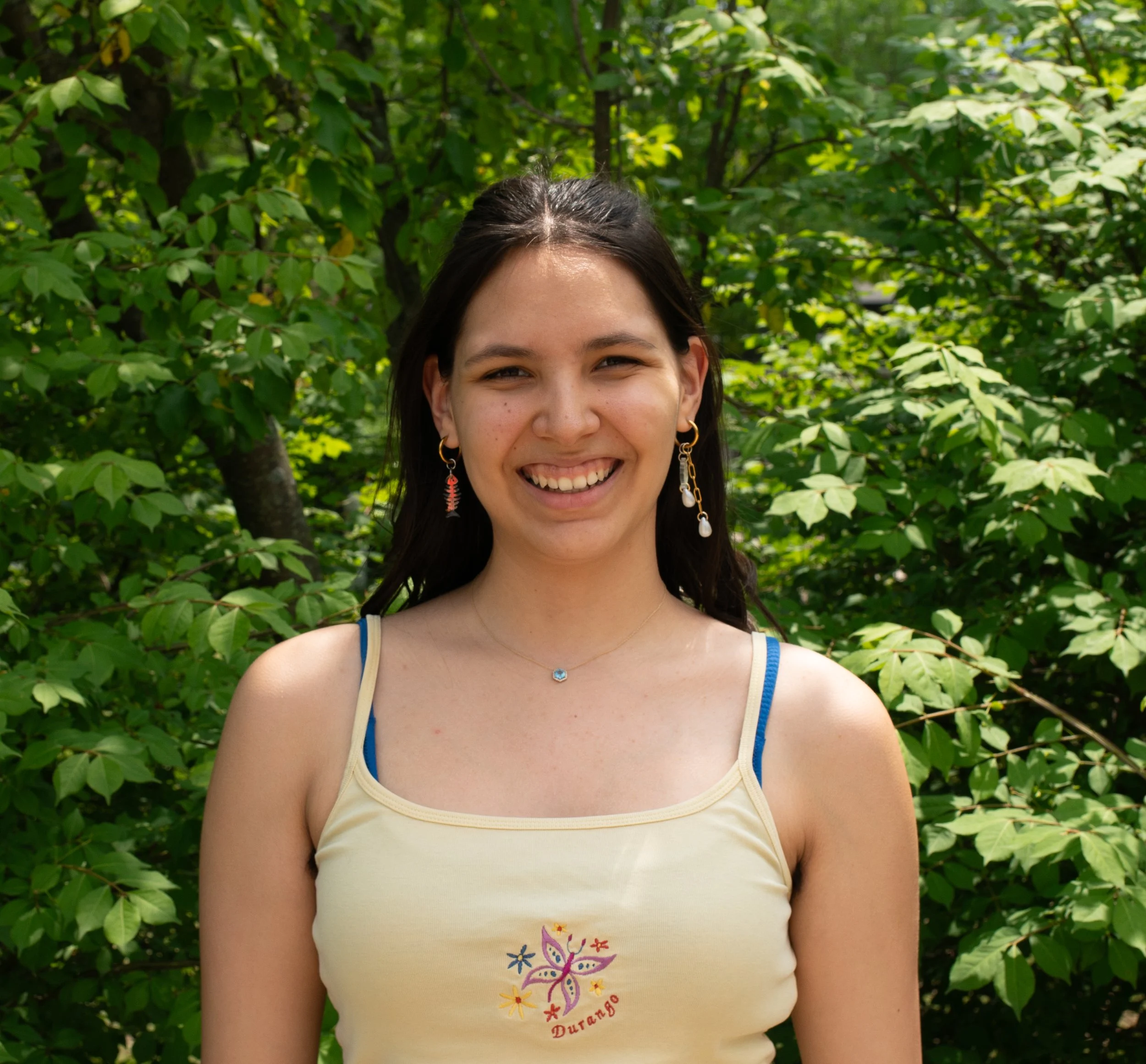 Young woman with curly dark hair wearing a gray and white striped t-shirt outdoors with green trees and blue sky in the background.