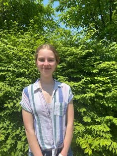 A young woman standing outdoors in front of lush green trees, smiling at the camera.