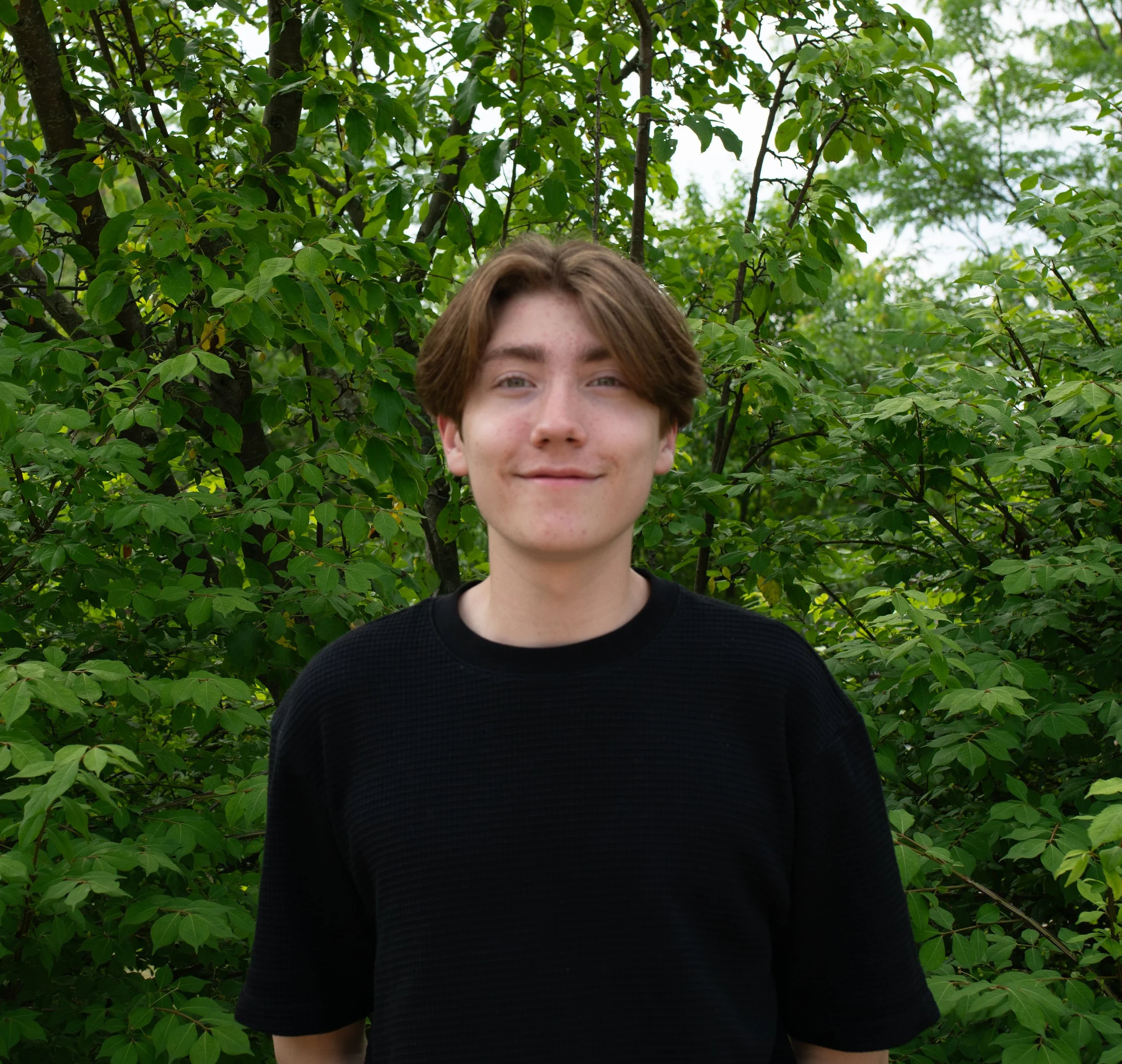 A young man with brown hair and glasses standing outdoors in front of green leafy bushes and trees on a sunny day.