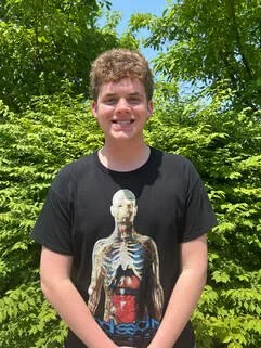 Young man standing outdoors in front of green trees, wearing a black T-shirt with a skeleton graphic.
