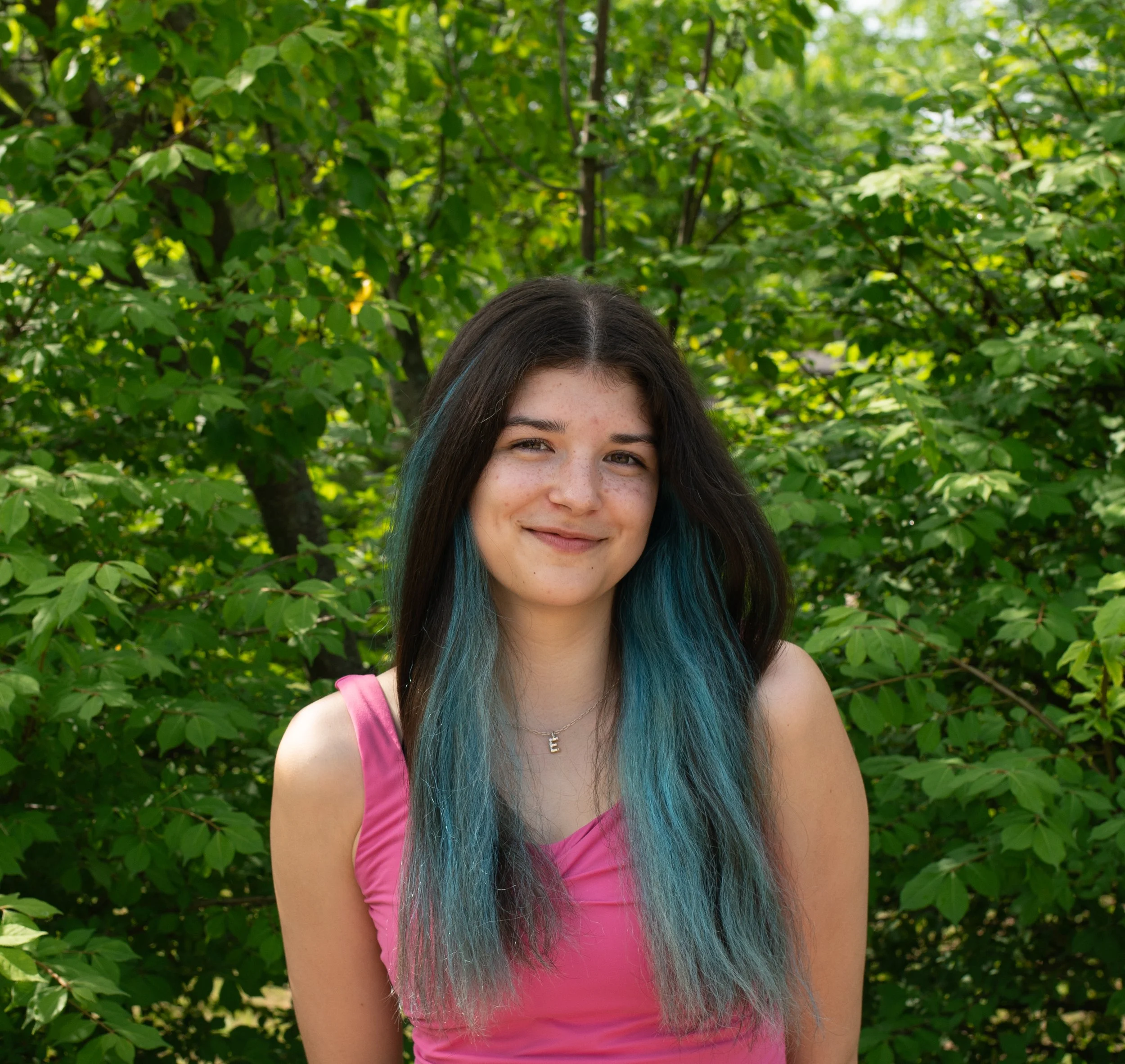 A young woman with short brown hair and blue eyes stands outdoors in front of lush green foliage, smiling with closed eyes. She is wearing a blue t-shirt and denim overalls with a chain belt, along with earrings and layered necklaces.