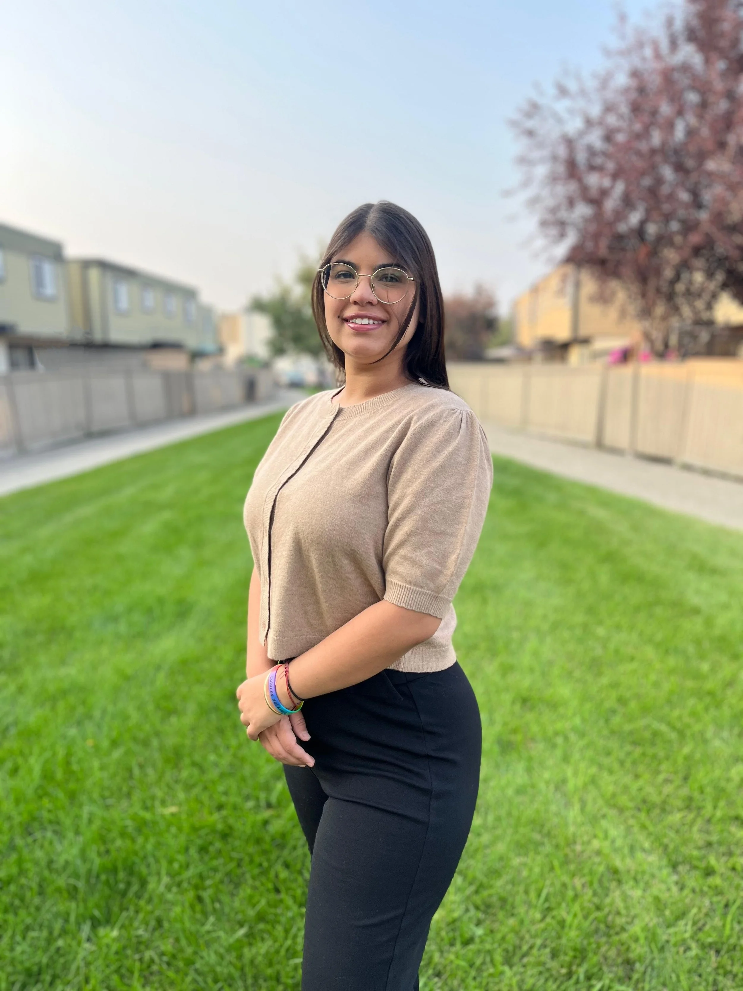 Young woman with glasses and dark hair standing on a grassy yard, smiling at the camera, with houses and trees in the background.