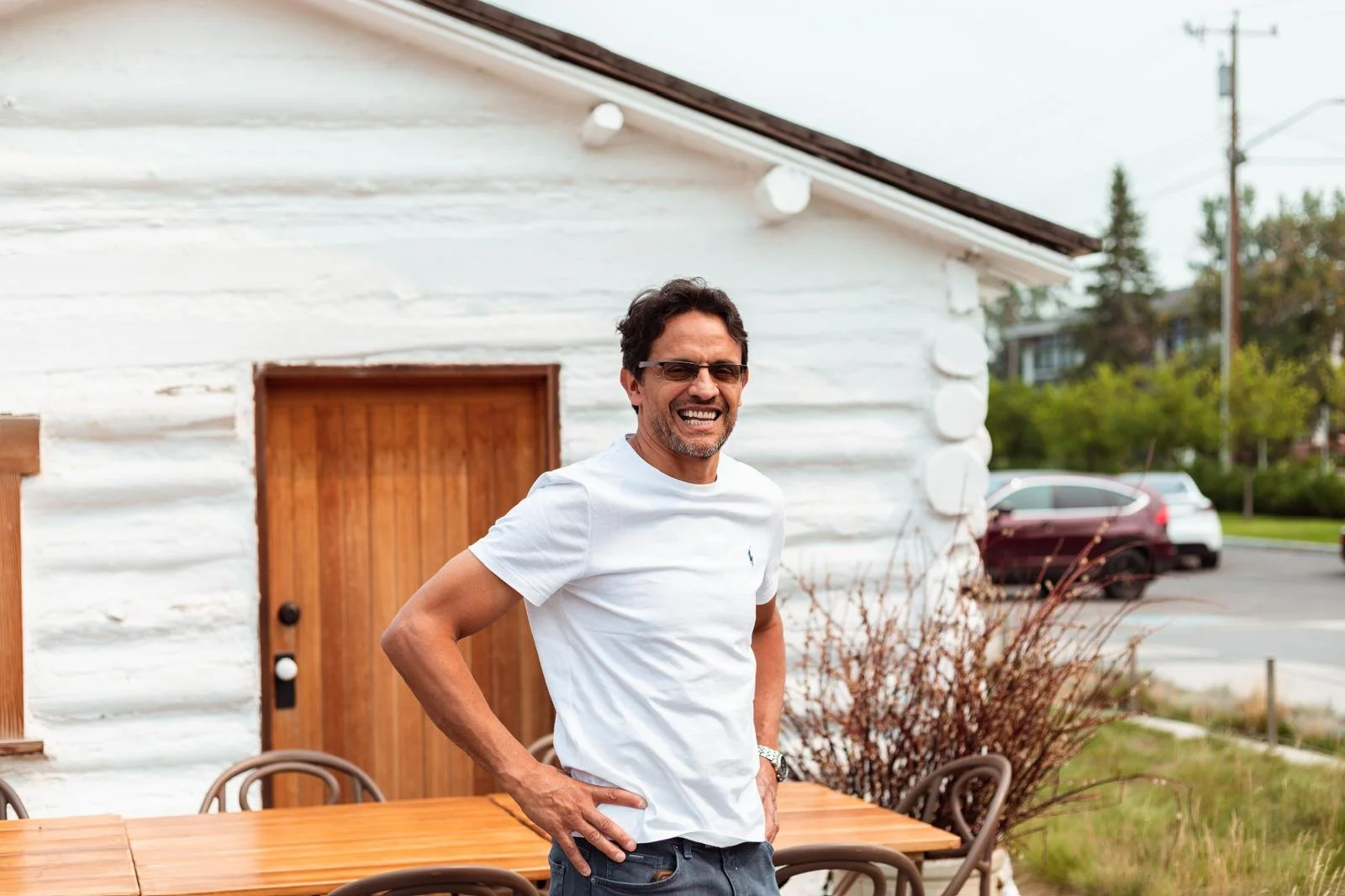 Smiling man in sunglasses wearing a white t-shirt standing outdoors beside a wooden table with chairs, with a white brick building and parked cars in the background.