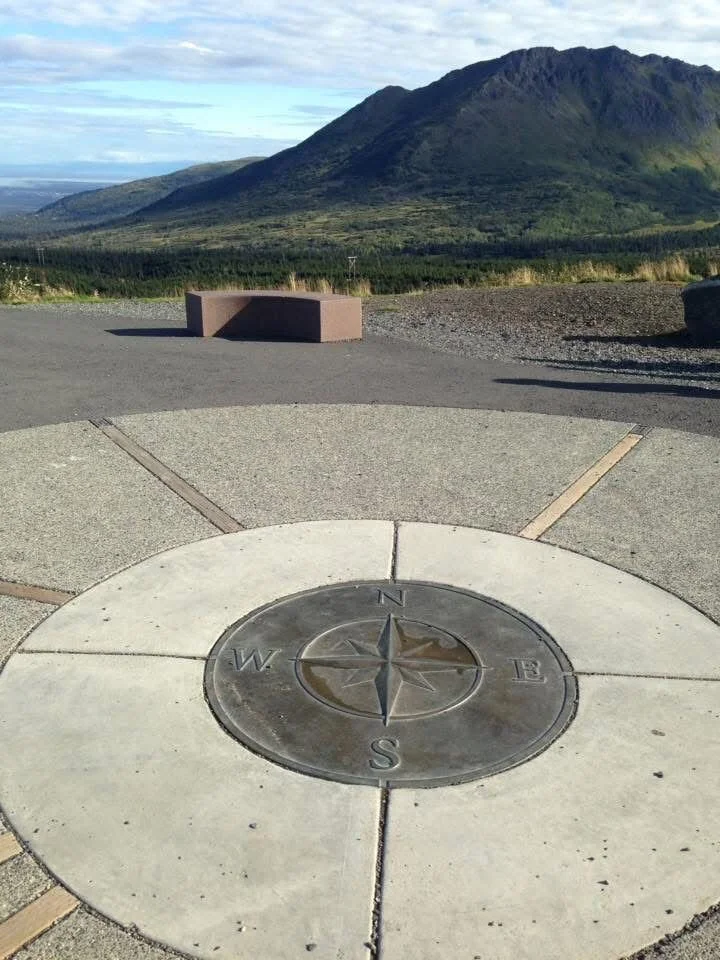 A compass rose embedded in the ground with a mountain and cloudy sky in the background.