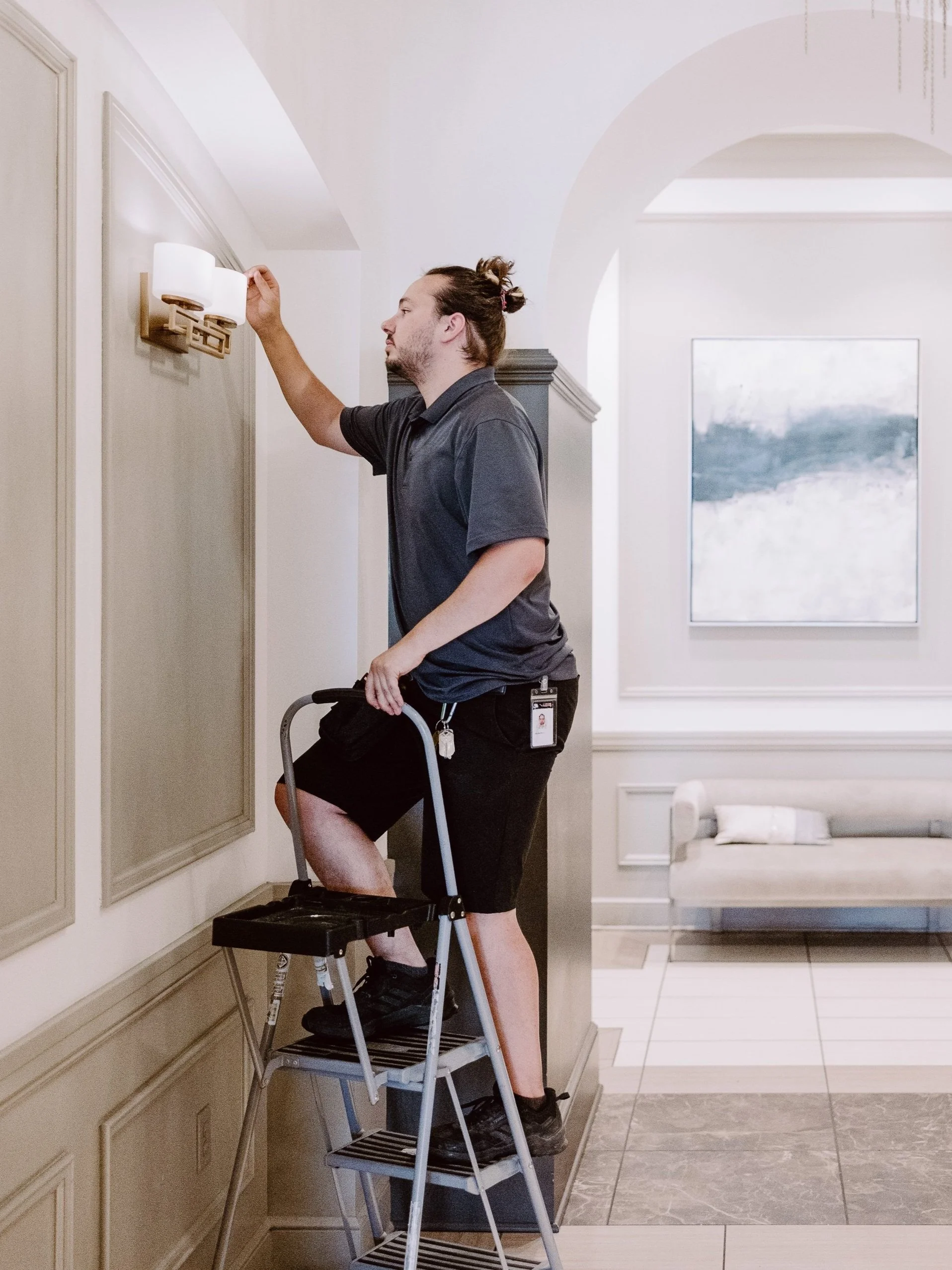 A man standing on a step ladder adjusting a wall-mounted light fixture in a modern interior space.