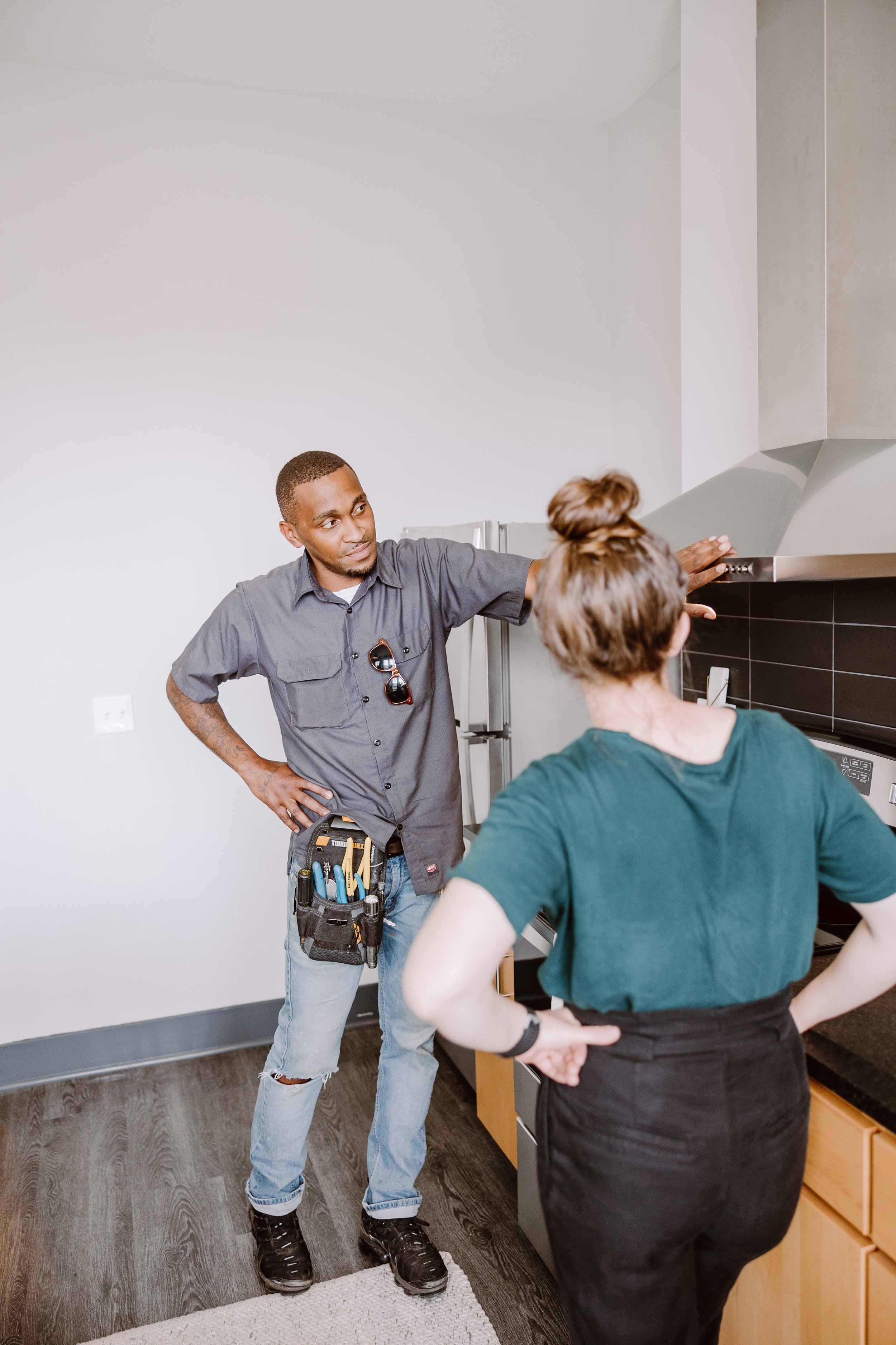 A man in a work uniform and tool belt discusses a kitchen appliance with a woman who has her back turned, inside a modern kitchen.