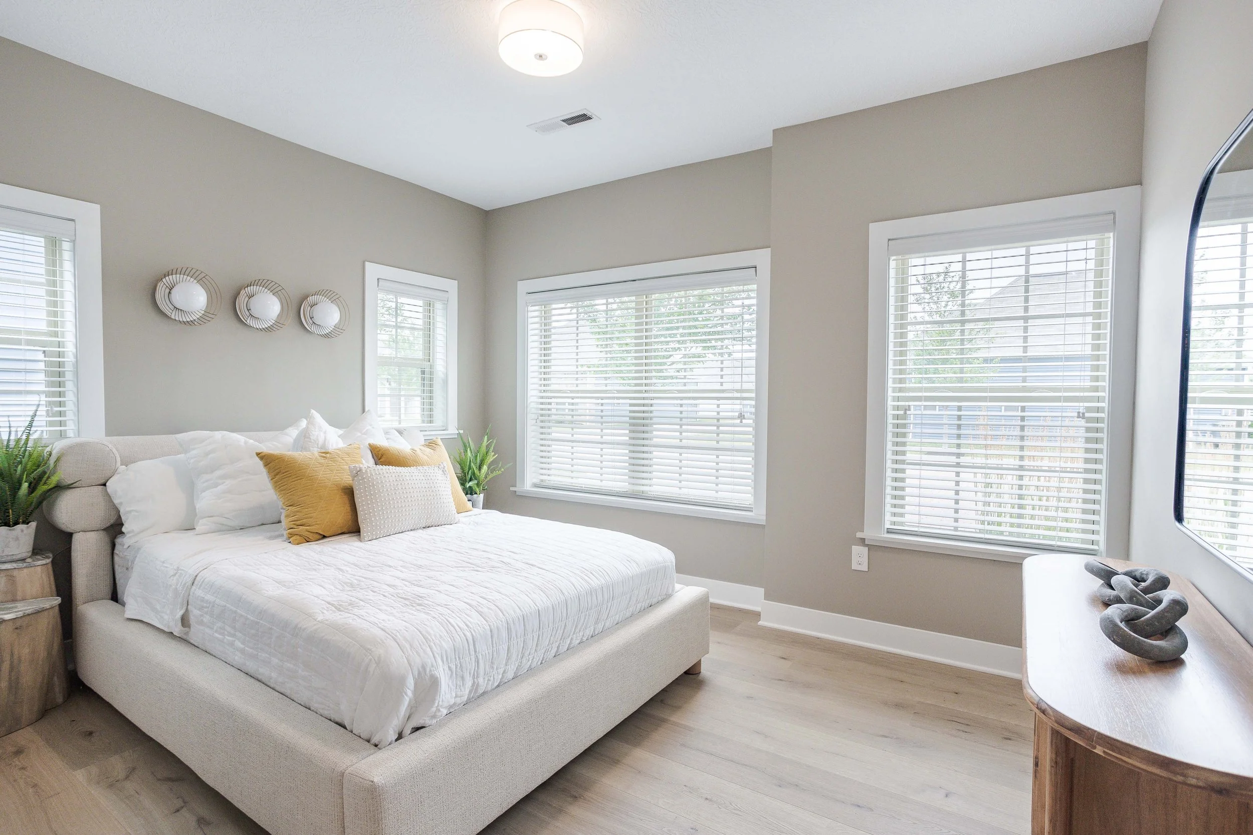 Bright bedroom with large window, neutral bedding, decorative pillows, and potted plants.