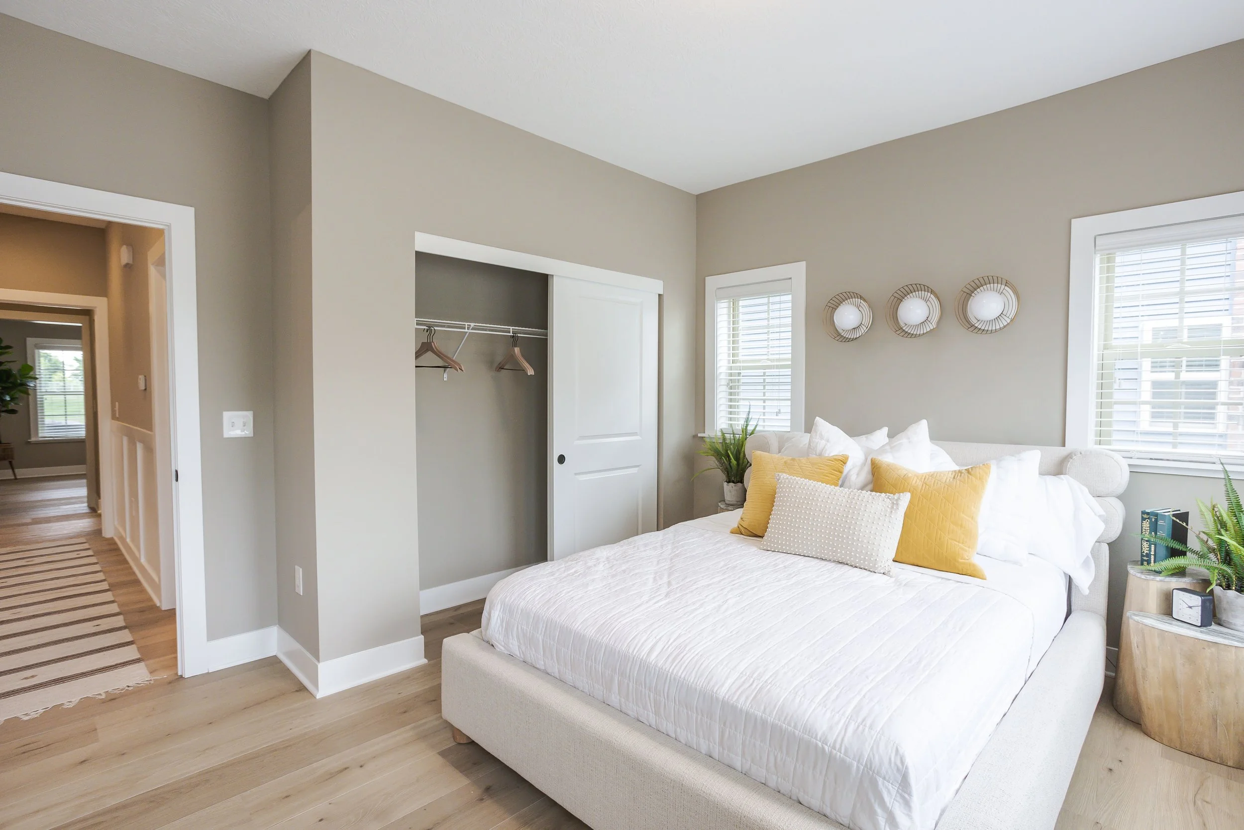 A bedroom with a cozy bed decorated with white and yellow pillows, two windows with blinds, a round wooden bedside table with plants and a clock, a small closet with sliding door, and a hallway visible through an open doorway.