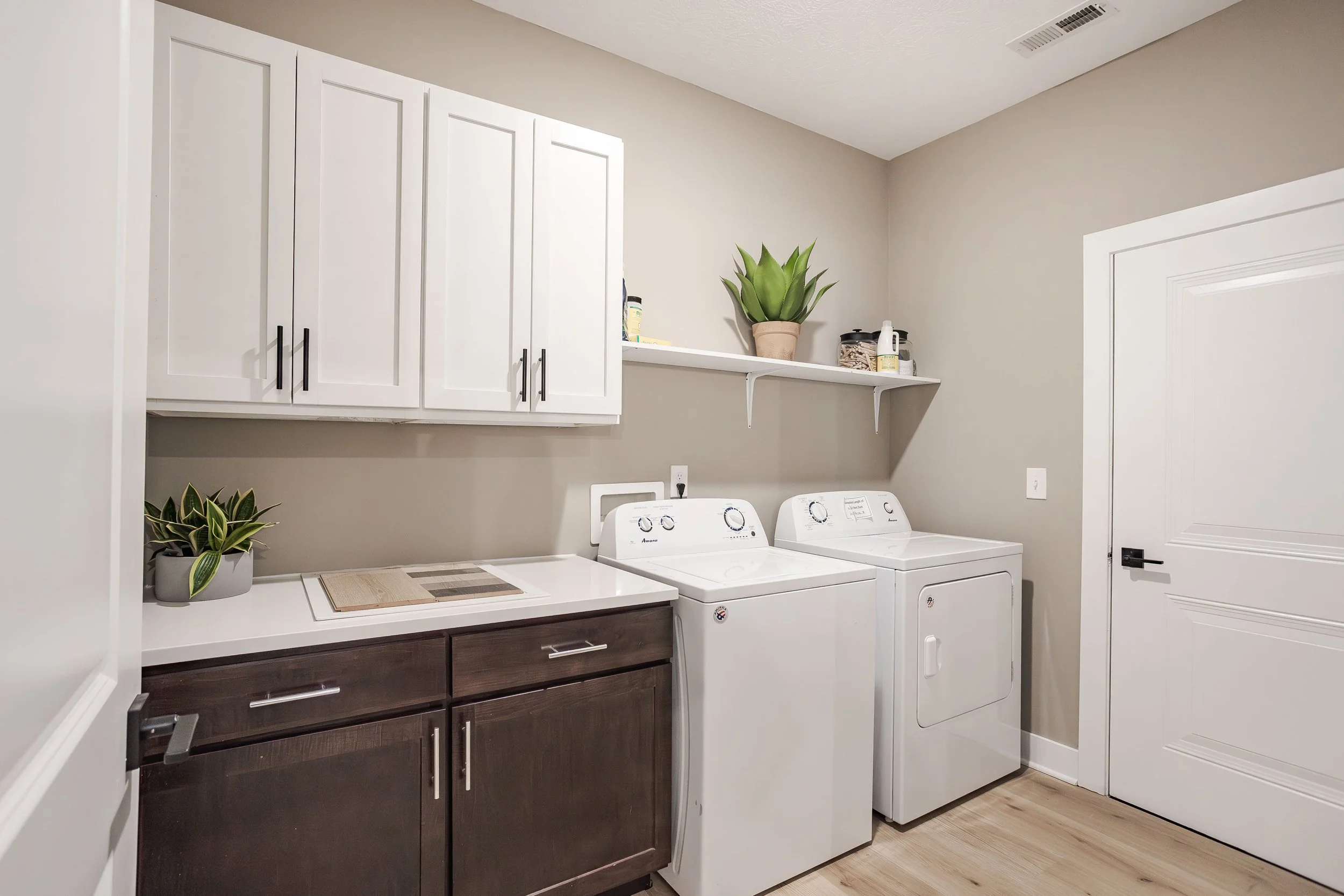 Laundry room with white cabinets, a shelf holding potted plants and laundry supplies, a white sink, a washing machine, and a dryer, with beige walls and light wood flooring.