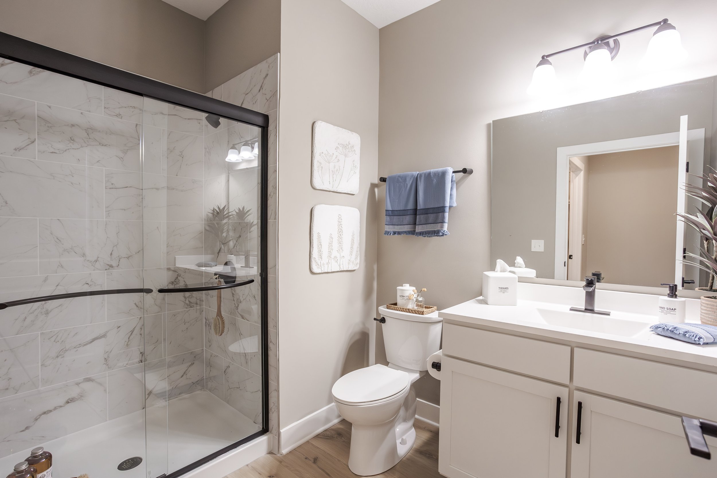 Modern bathroom with a glass-enclosed shower, white vanity with a large mirror, toilet, and decorative wall art; featuring neutral tones and minimalist decor.