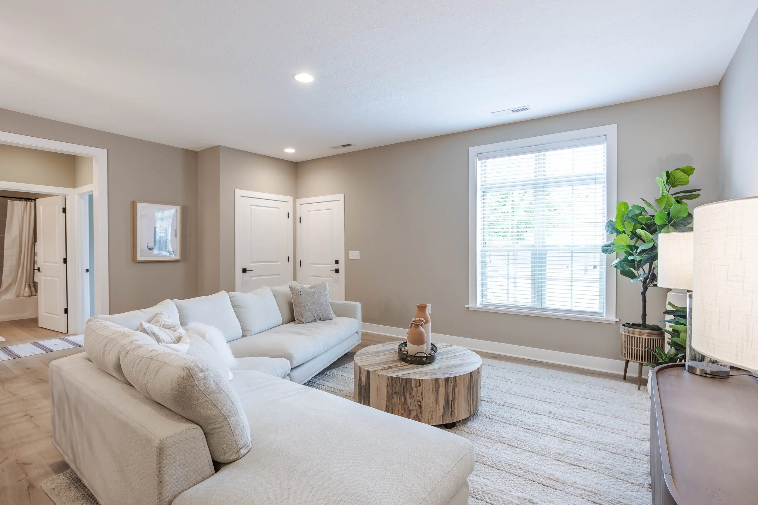 Living room with a white sectional sofa, a round wooden coffee table, a large window with blinds, a potted plant, and a lamp.