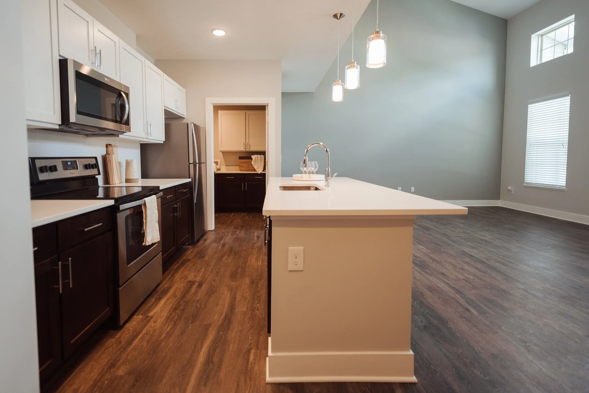 Open-concept kitchen with white upper cabinets, dark lower cabinets, stainless steel appliances, a kitchen island with a sink, pendant lights, and a large living area with hardwood floors and windows.