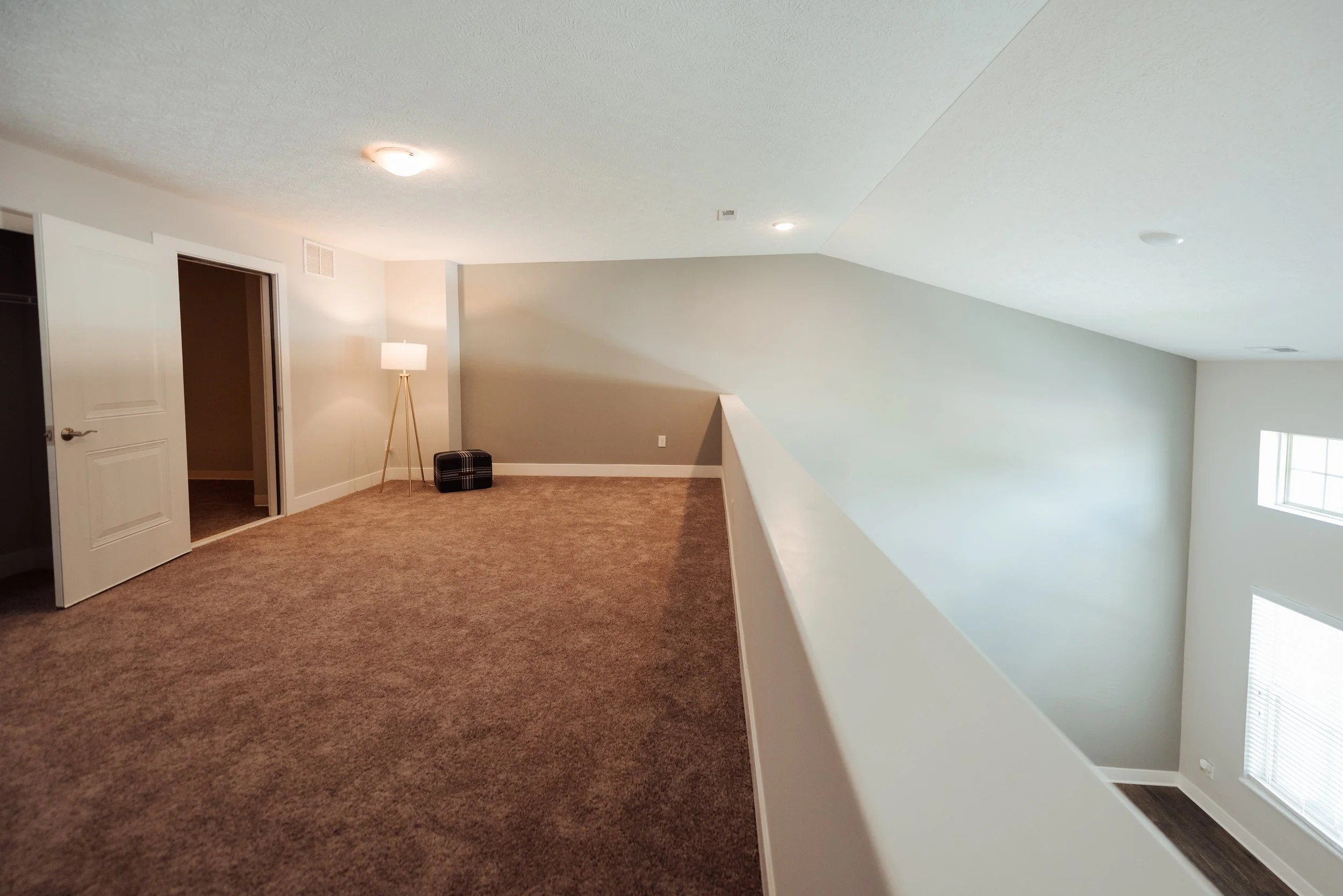 An empty upper floor of a house with beige carpet, a floor lamp, and two closed closet doors, overlooking a lower level with large windows.