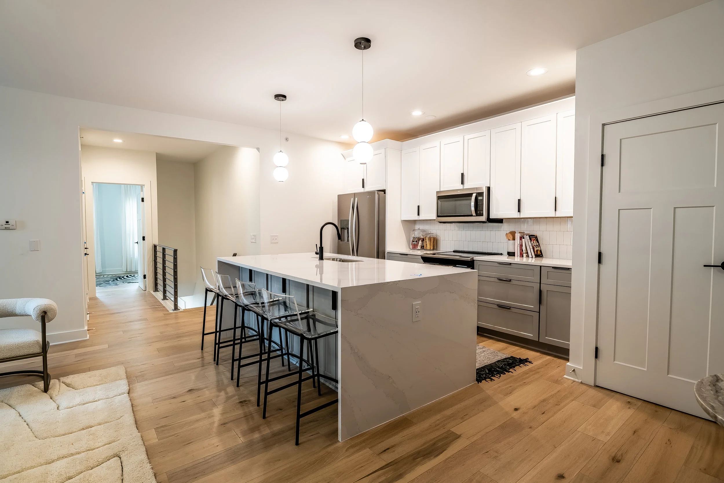 Modern kitchen with white upper cabinets, grey lower cabinets, stainless steel refrigerator, microwave, and oven. An island with a white marble countertop, three transparent barstools, and pendant lighting.
