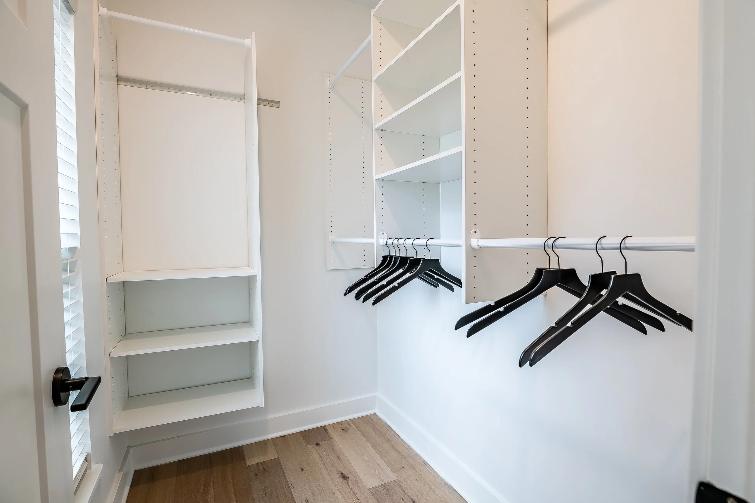 Empty white walk-in closet with black hangers on white rods and built-in shelves, hardwood floor, and a partially visible window with blinds.