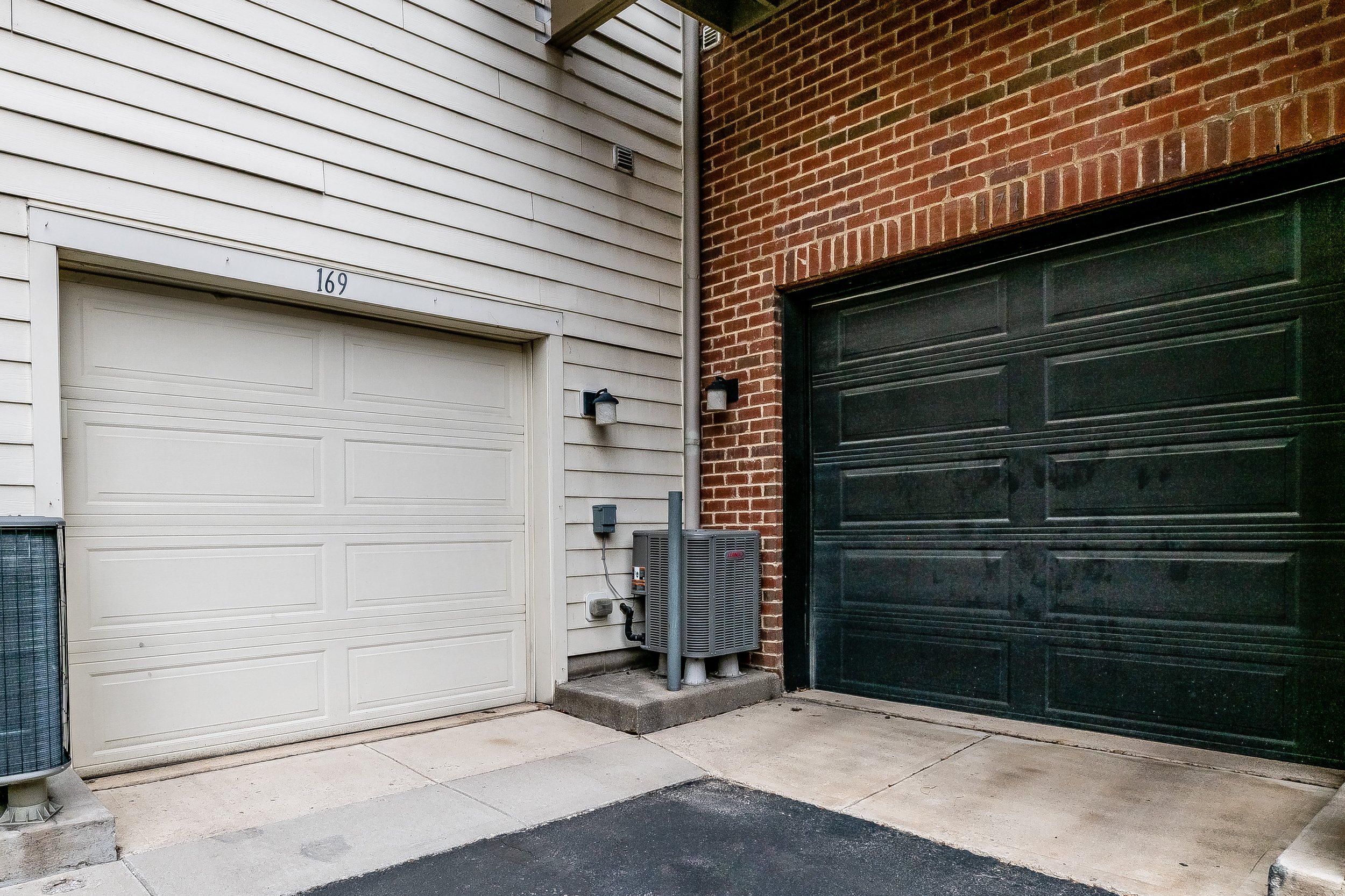 Two garage doors, one white and one black, with brick and siding walls, an air conditioning unit, and outdoor lights.