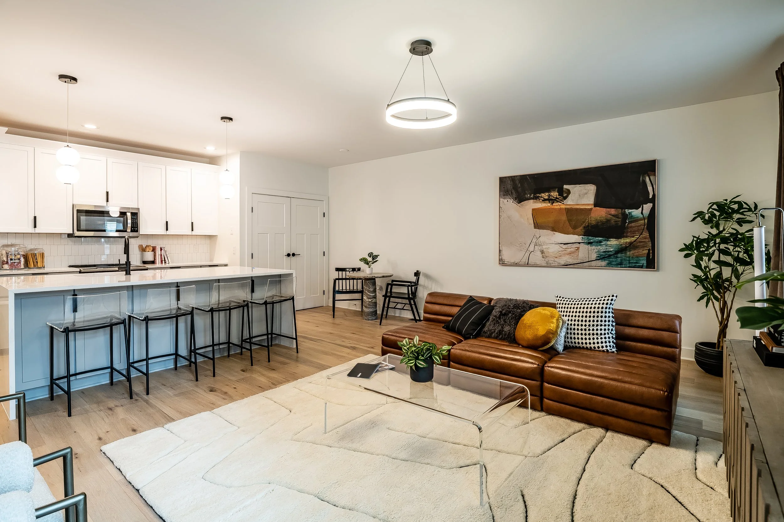 Modern open-concept living room and kitchen with white cabinets, black bar stools, leather sofa with pillows, abstract wall art, potted plants, and contemporary lighting fixtures.