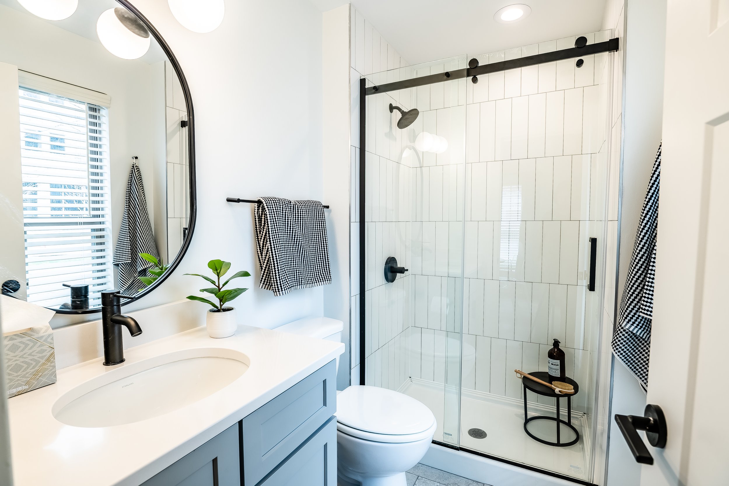 Modern bathroom with white walls and a glass-enclosed shower with white tiles, a black showerhead, and black fixtures. There is a white vanity with a black faucet, a round mirror, a potted plant, and black and white checkered towels.