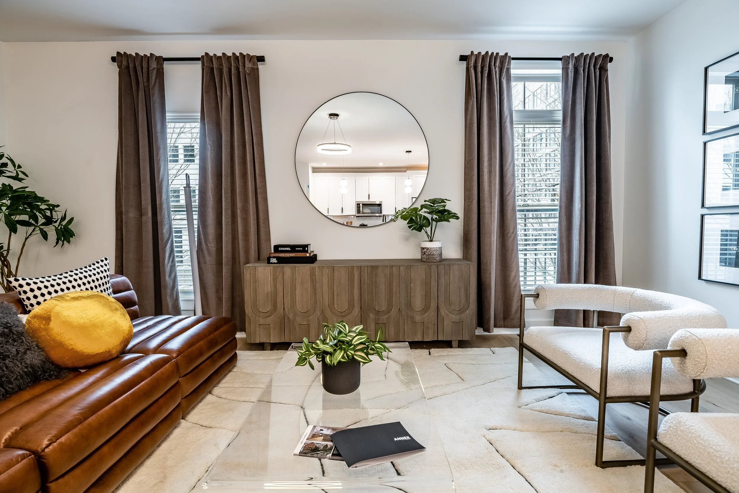 Modern living room with brown leather sofa, white armchairs, a glass coffee table, potted plants, large window with brown curtains, round mirror on the wall, and a wooden sideboard.