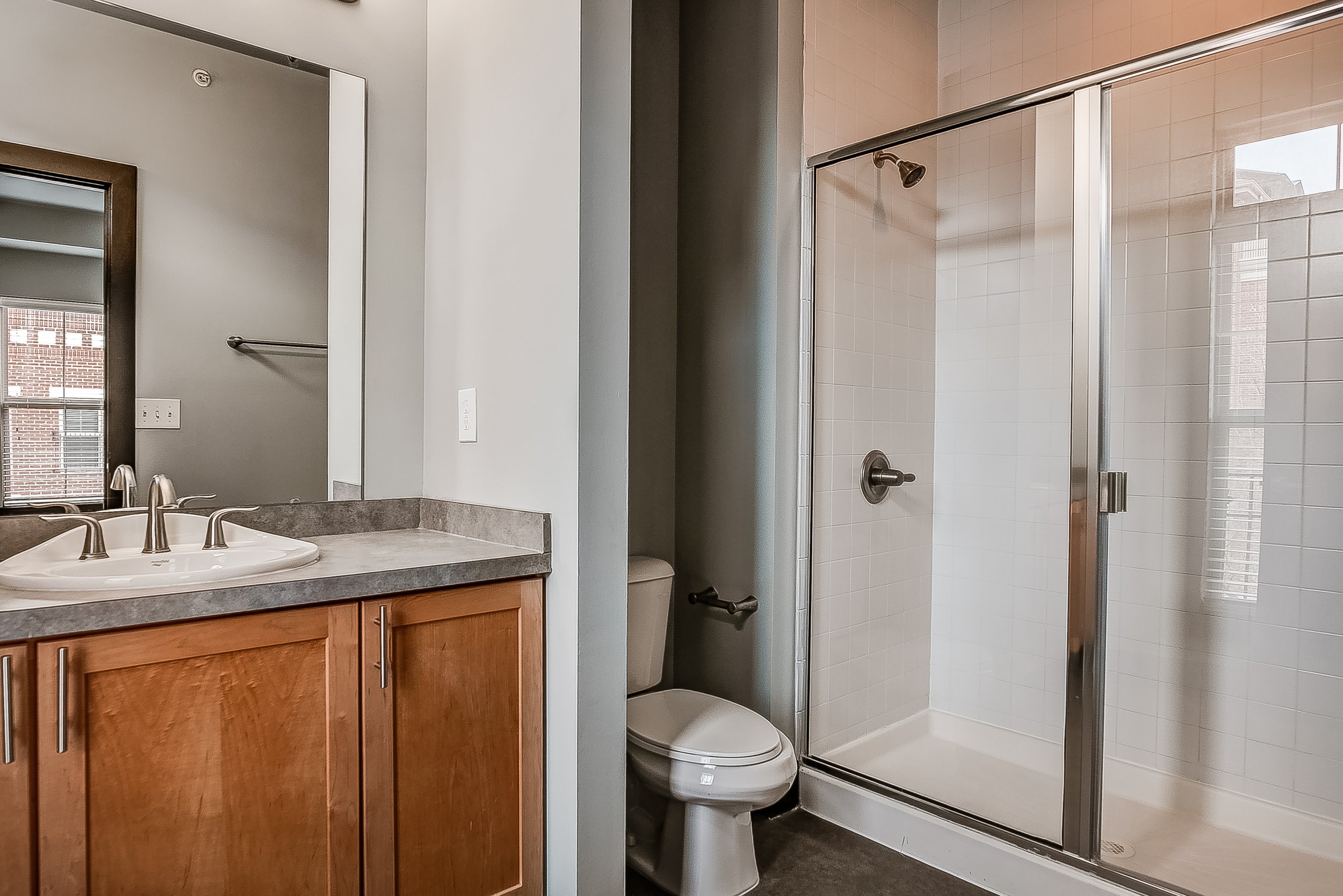 Bathroom with wooden vanity, gray countertop, white sink, and silver faucet; mirror and window above vanity; toilet next to shower with glass door and white tiled walls.
