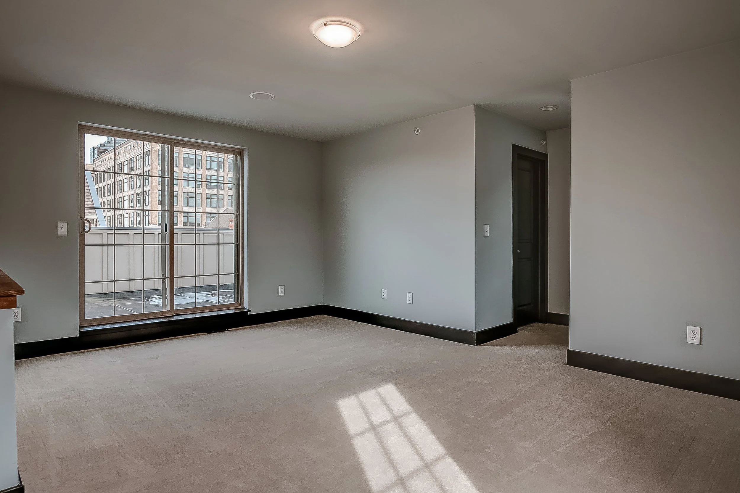 Empty living room with beige carpet, gray-painted walls, black baseboards, a sliding glass door leading to a small balcony with urban buildings outside, and a ceiling light fixture.