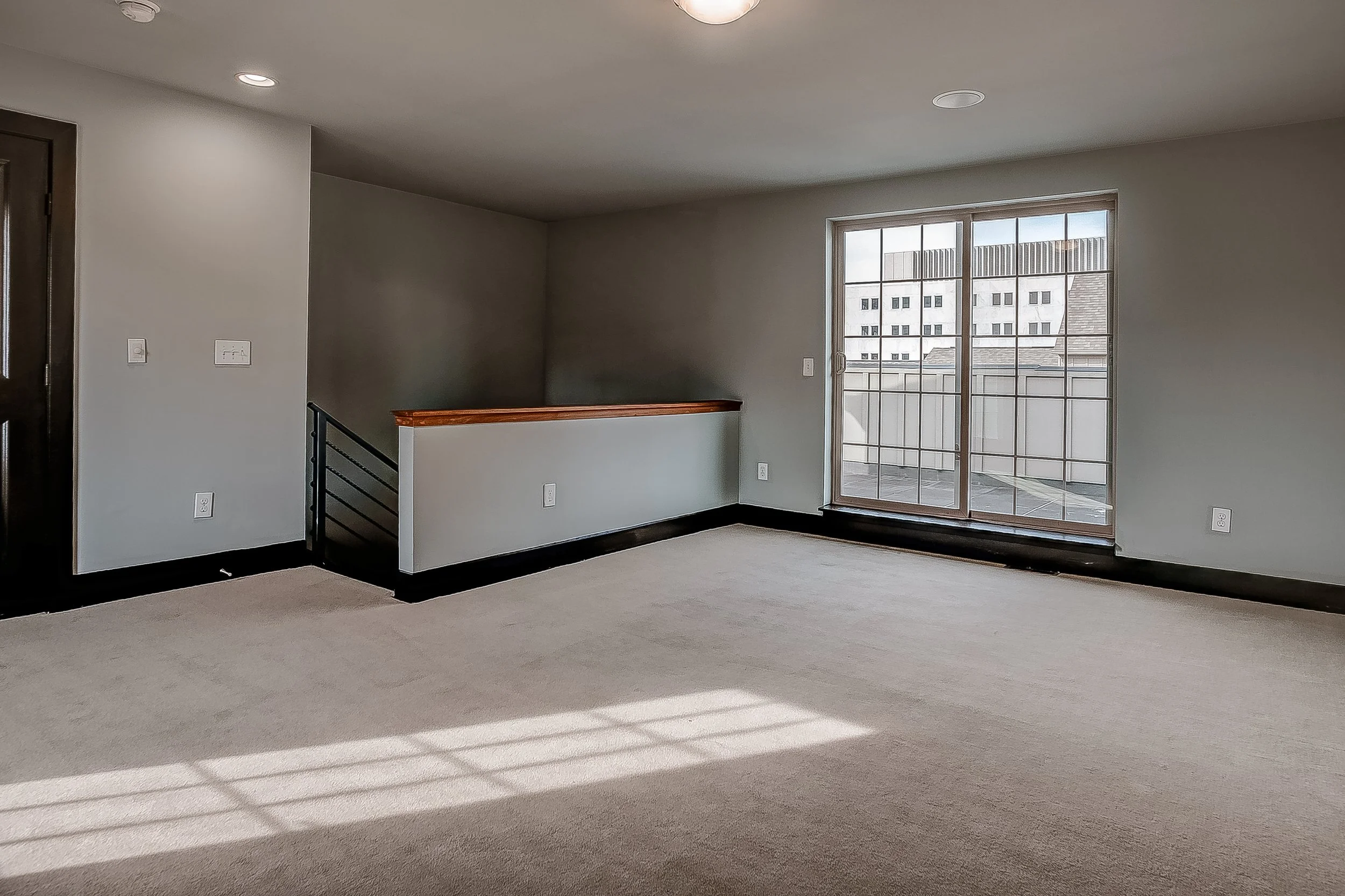 Empty living room with beige carpet, gray walls, black baseboards, a sliding glass door leading to a balcony, and city buildings visible outside.