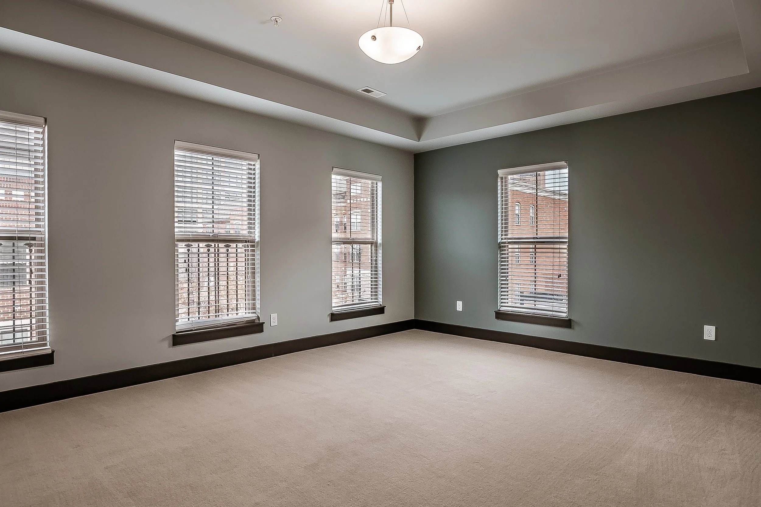 Empty room with four windows, beige carpet, gray wall, white ceiling with a ceiling light, and black baseboards.